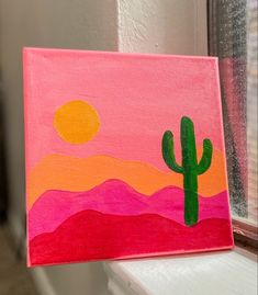 a painting on a window sill with a cactus in the foreground and a sunset behind it