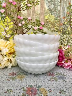 four white bowls sitting on top of a table next to colorful flowers and trees in the background