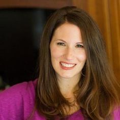 a woman with long brown hair smiling at the camera and wearing a purple shirt in front of a wood paneled wall