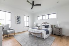 a bedroom with white walls and wood flooring has a bed, two chairs, and a ceiling fan