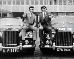 two men in suits sitting on the front of an old car with their thumbs up