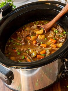 a crock pot filled with beef stew and carrots next to a wooden spoon