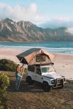 a woman standing under an umbrella next to a truck on the beach with mountains in the background