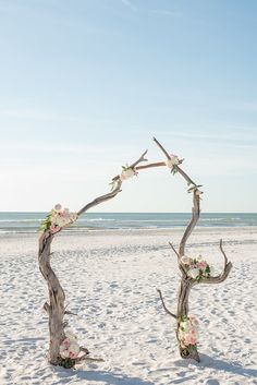 an arch made out of driftwood on the beach is decorated with flowers and greenery