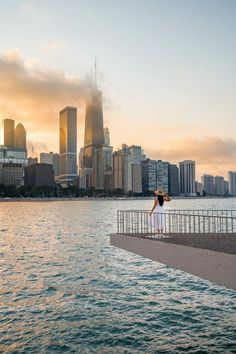 a woman standing on the edge of a pier looking out at the water and skyscrapers in the background