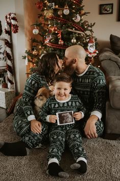 a man and woman are sitting in front of a christmas tree with a baby on their lap