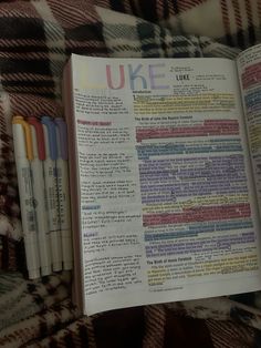 an open book sitting on top of a plaid blanket next to pens and marker pens