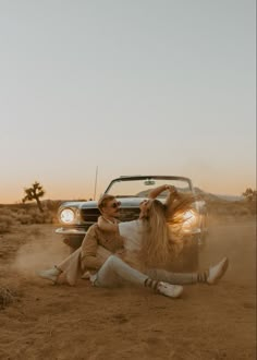 a man and woman sitting on the ground next to a car in the desert at sunset