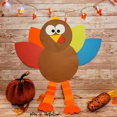 a paper turkey is standing next to a pumpkin and some string lights on a wooden wall