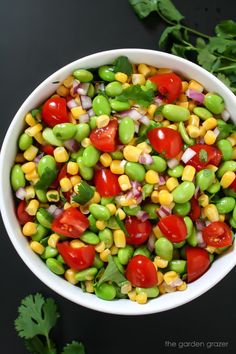 a white bowl filled with corn, tomatoes and green beans next to cilantro