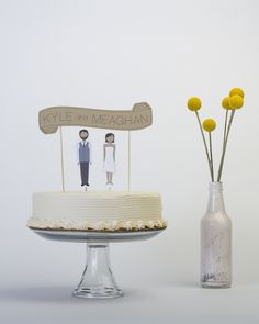 there is a cake with two people on it and flowers in the vase next to it