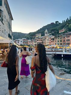 two women walking down the street in front of some buildings with boats parked on the water