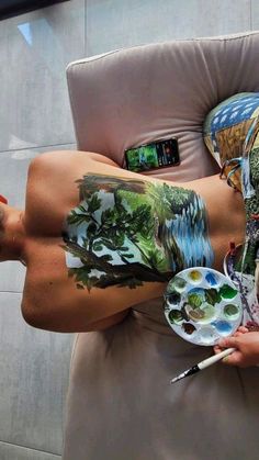 a man with tattoos on his stomach holding a paintbrush and palette while laying in a chair