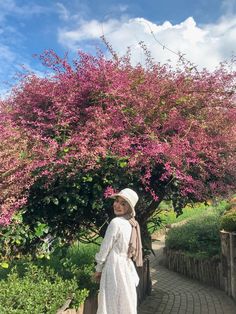 a woman in a white dress and hat standing under a tree with pink flowers on it