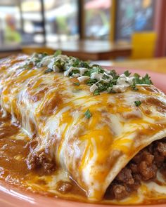 a pink plate topped with a burrito covered in cheese and meat, garnished with green onions