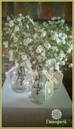 two clear vases with white flowers in them on a table next to a brick wall