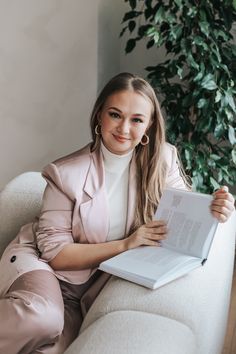 a woman is sitting on a couch holding a book and smiling at the camera while wearing a pink suit