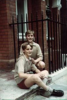 two young men sitting on the sidewalk in front of a fence and gate, one with his arm around the other's neck