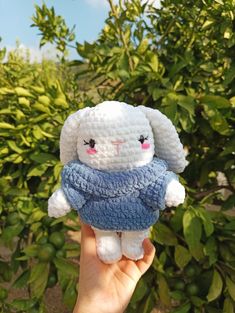 a hand holding a small white stuffed animal in front of some green bushes and trees