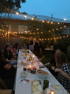 a group of people sitting at a long table with food on it and lights strung over them