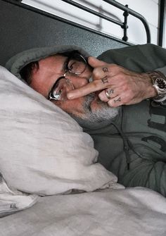 a man in glasses laying in bed with his hand up to his face, making the peace sign