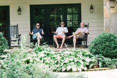 three people sitting on chairs in front of a house with plants and bushes around them