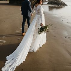 a bride and groom walking on the beach holding each other's hands while they hold flowers