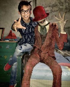 two men dressed up like clowns sitting on top of a bed with their hands in the air