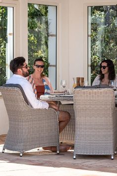 three people sitting at an outdoor table with wine glasses on it and one person standing next to the table