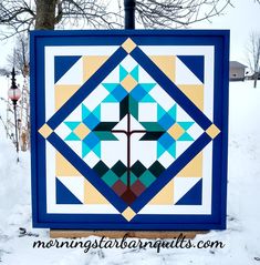 a blue and yellow quilt hanging on the side of a building in the snow with trees behind it