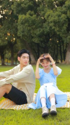 a young man and woman sitting on a blanket in the grass with trees behind them