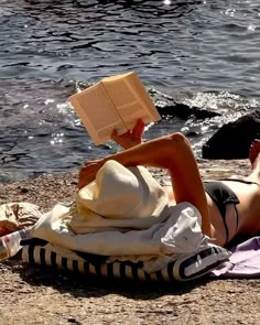 a woman reading a book while laying on the ground next to some water and rocks