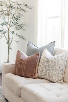 a white couch with several pillows on it in front of a potted plant and window