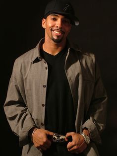 a man in a black shirt and hat smiles while standing with his hands in his pockets