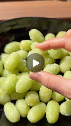 a hand is holding a green object in a black bowl filled with grapes and water