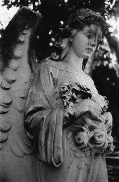 black and white photograph of an angel holding a flower arrangement in her hand, with trees in the background