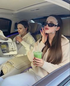 two women sitting in the back seat of a car, one holding a drink while the other holds up her hand