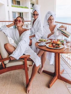 three women in white robes sitting at a table with food and drinks on the balcony