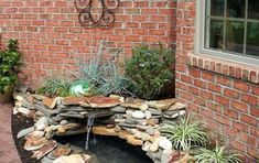 a small pond in front of a brick building with plants and rocks on the ground