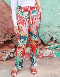 a woman standing in front of a green wall wearing colorful pants and white shirt with palm leaves on it