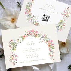 two wedding cards with flowers on them next to some white ribbons and flowers in the background