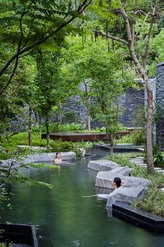 a man is sitting in the water next to some rocks and trees, while another person looks on