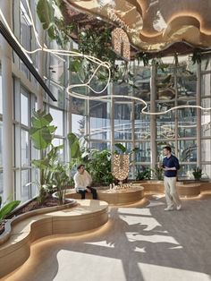 two people are sitting in the sunlit room with large windows and potted plants