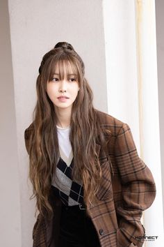 a woman with long hair standing next to a white wall wearing a brown jacket and black pants