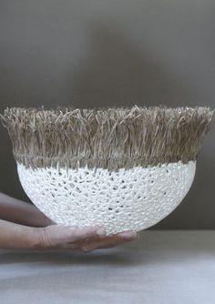 a hand holding a white and brown bowl with grass in it's center, on top of a table