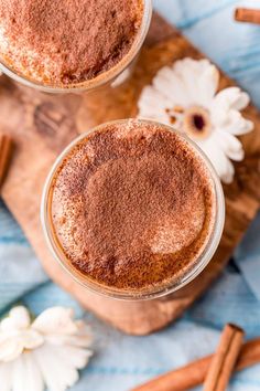 two glasses filled with cinnamon drink on top of a wooden cutting board next to flowers