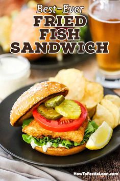 a fish sandwich with pickles and tomatoes on a black plate next to potato chips