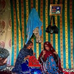 two women sitting on the floor in front of a wall covered with colorful tapes and cloths