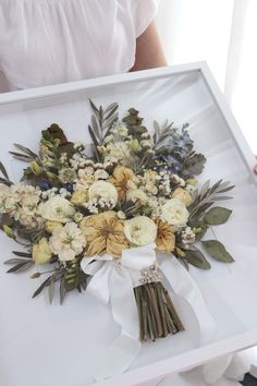 a bouquet of flowers is displayed in a white frame on the table next to a woman's hand