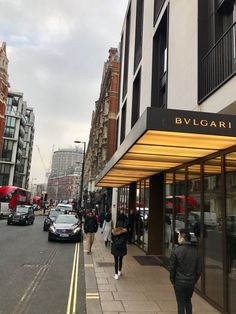 people are walking down the sidewalk in front of a building on a busy city street